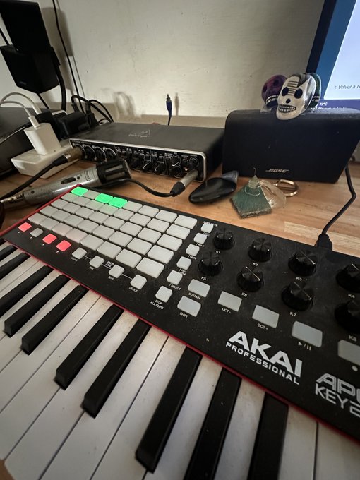 A music production setup featuring an AKAI Professional keyboard controller with piano keys and pads, along with an audio interface and a Bose speaker on a desk.