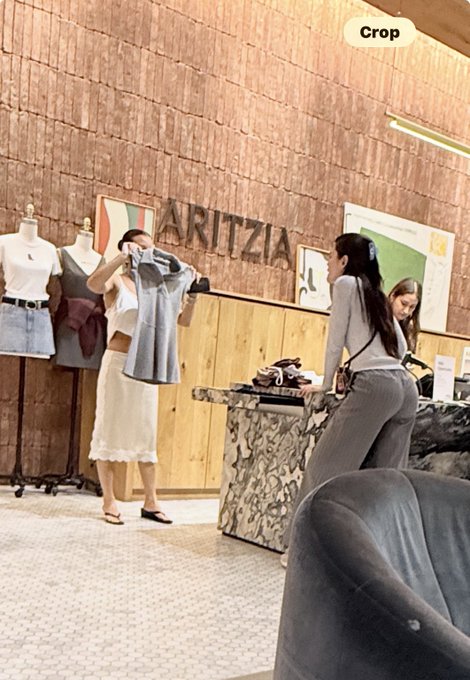 A woman tries on a gray top inside an Aritzia store while another woman in a fitted outfit looks on near a marble counter, capturing the moment of a fashion verdict as the shopper declares "The bottom is in. I've seen enough."