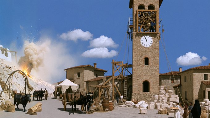 A historical or fantasy scene showing a stone clock tower in a village setting, with an explosion occurring in the background near horse-drawn carts and figures in period clothing.