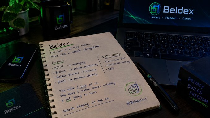 A notebook page on a desk displaying handwritten notes about 'Beldex' as a privacy token, surrounded by Beldex-branded items including a laptop, pen, and mug, with the text 'Privacy Builds Freedom' visible.