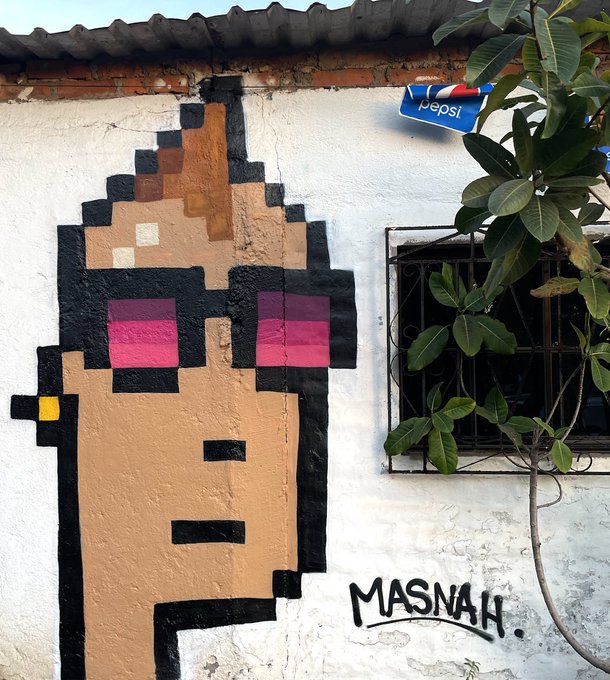 Street art mural painted on a white wall depicting a pixelated character with pink sunglasses, signed 'MASNAH' with a Pepsi sign and plant nearby.