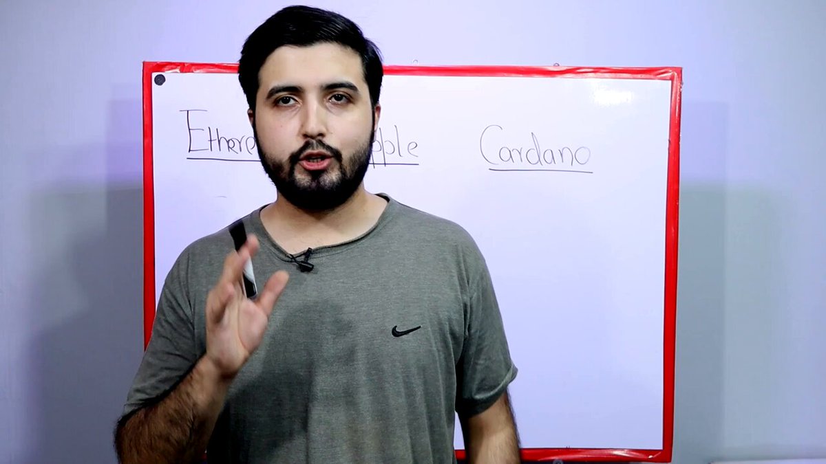 The image shows a man wearing a gray t-shirt standing in front of a whiteboard with the words "Cardano" written on it, along with some text. The man appears to be giving a presentation or explanation about Cardano, possibly discussing its features and benefits.
