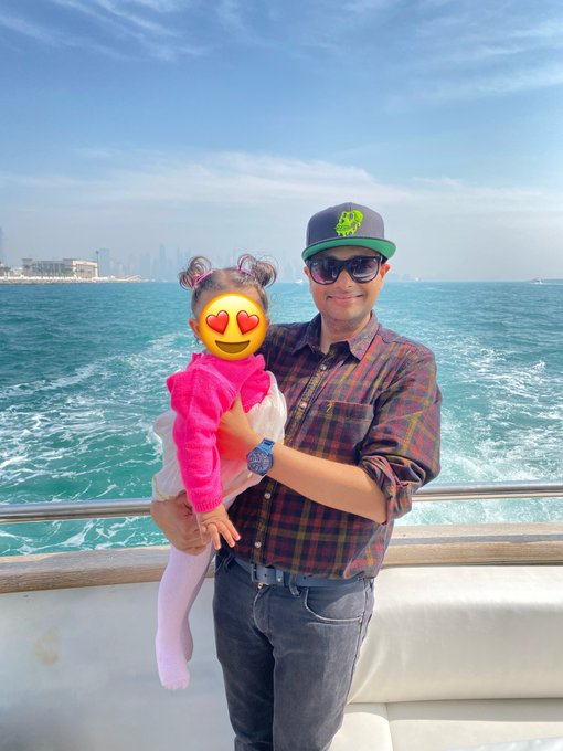 A man wearing a cap and sunglasses holds a young child in a pink sweater on a boat, with the sea and a city skyline behind them.