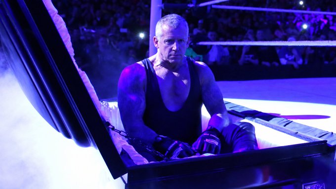 A muscular wrestler in a black tank top sits inside a casket at a wrestling arena, bathed in purple lighting with smoke effects and a crowd visible in the background.