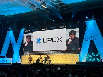 A cryptocurrency conference with 'UPCX' and 'CTRL' branding on stage screens, showing speakers and seated presenters under blue stage lighting