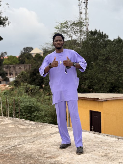A young man wearing a light purple traditional outfit poses on a rooftop, pointing toward the camera.