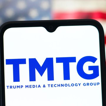 The image shows the Trump Media & Technology Group (TMTG) logo displayed on a smartphone screen with a blurred American flag in the background, illustrating the company behind the significant Bitcoin sales mentioned in the post.