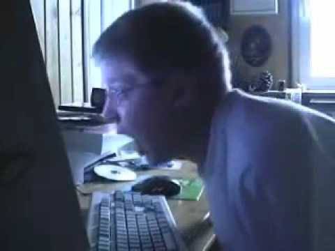A grainy photograph of an older man with gray hair and glasses wearing a light-colored shirt, sitting at a desk and looking at a computer monitor with a keyboard in front of him.