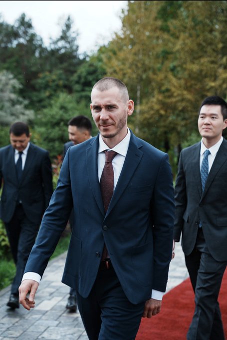 A photograph of a man in a dark suit and brown tie walking on a red carpet, flanked by other suited men in an outdoor setting with trees.