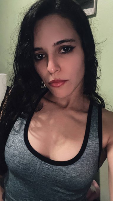 A selfie of a young woman with long dark hair wearing a gray tank top with black trim, looking directly at the camera.
