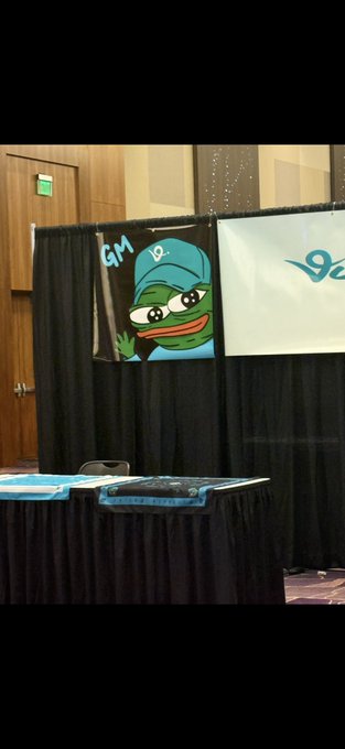 A booth setup at what appears to be a convention, featuring a Pepe the Frog character wearing a cap with 'GM' and '19' visible, along with other banners.