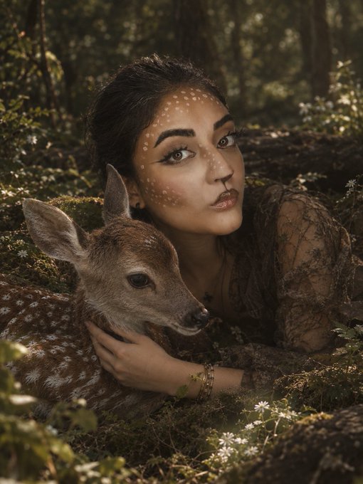 Portrait-style photo of a woman with deer-like face paint and makeup posing closely with a young fawn in a forest setting.