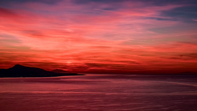A dramatic sunset paints the sky in vibrant shades of red, orange, and purple over a calm body of water, with a dark landmass silhouetted on the left and a contrail streaking across the sky.