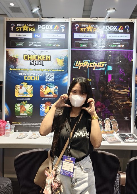 A masked individual poses with a heart gesture in front of the "Chicken Saga" and "Uprising Legends" indie game booths at PGDX (Philippine Game Dev Expo) 2023. The accompanying post says Reed is a core moderator for @Kusho_World, a new metaverse and blockchain gaming platform.