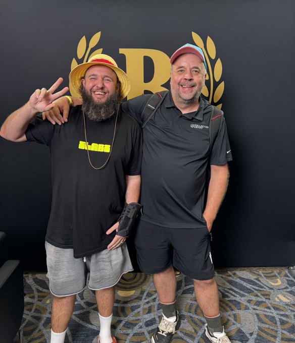 Two smiling men pose together in front of a gold "BC" (likely Bitcoin Conference) logo backdrop, with one wearing a yellow straw hat and flashing a peace sign while the other sports a red cap and a black polo with a "Bitcoin" logo. The image likely commemorates a fan meeting with a notable Bitcoin figure, as suggested by the caption praising "Tuom" and showing appreciation to "Collector Crypt."