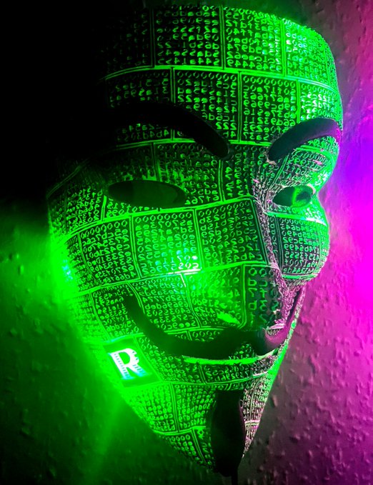 A Guy Fawkes-style mask covered in projected green digital code or binary patterns, lit with green and purple neon lighting.