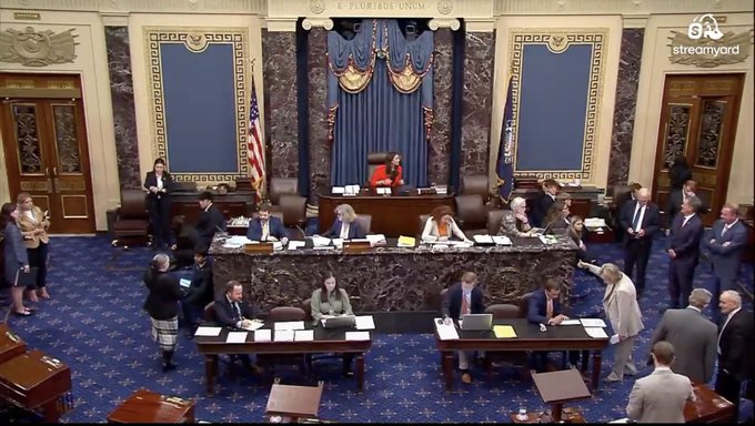 A wide shot of the US Senate chamber during a session, featuring the presiding officer, clerks, and various senators and staff at their desks.