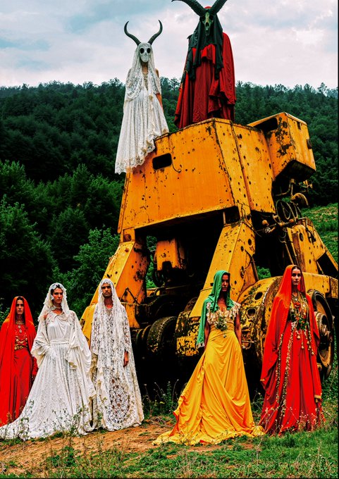 A highly stylized fashion photograph featuring women in elaborate, brightly colored dresses and masked figures with horns standing on and around a large, rusty yellow piece of machinery.