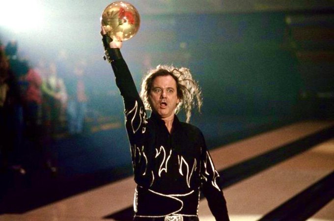 A man with wild, electrified hair wearing a black shirt with white flame-like designs triumphantly holds up a shiny golden disco ball above his head, striking a dramatic pose that suggests a victorious celebration in a dimly lit arena with a small crowd visible in the background.