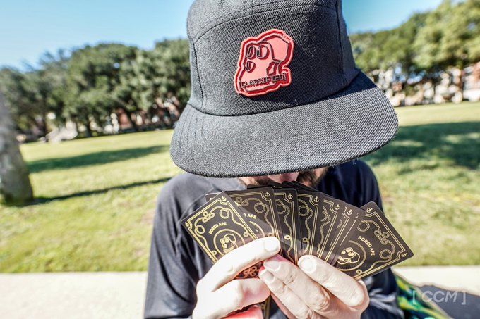 An outdoor portrait shows a person in a dark bucket hat holding a fan of black-and-gold cards; the hat patch includes the word “CLASSIFIED,” and a small watermark is visible at bottom right.