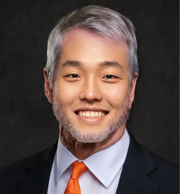 The image features a man wearing a suit, tie, and white shirt with gray hair. He has an orange tie that stands out against his dark-colored attire. The background of the photo appears to be black, which contrasts with the man's light-colored hair and highlights him in the frame. The caption "Steady lads $BTC" suggests a connection between the image and a post about investing or financial matters.