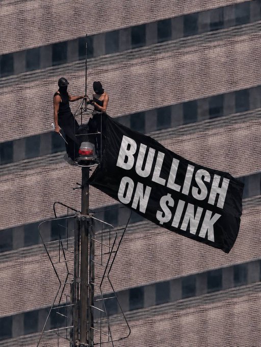 Two people in black clothing and ski masks sit atop a flagpole on a tall building, deploying a black banner reading 'BULLISH ON $INK'.