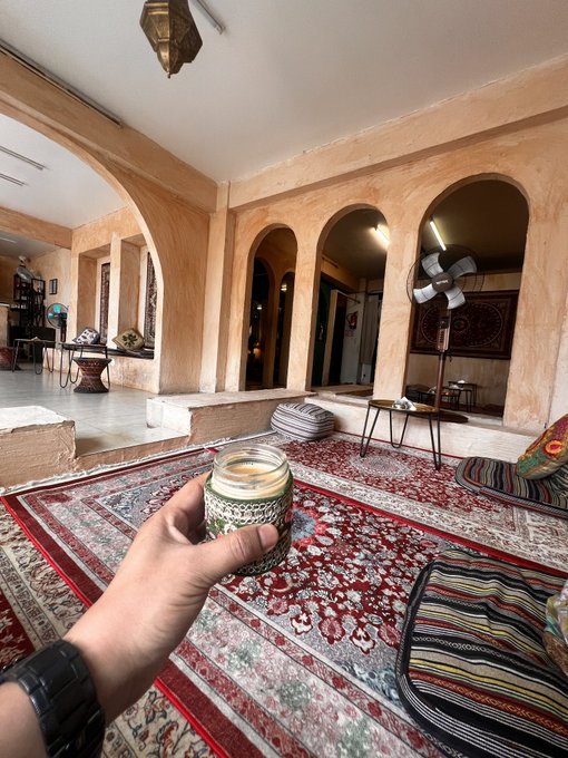 A first-person perspective photo of a hand holding a decorated ceramic mug on a patterned Persian rug, with traditional Middle Eastern architecture featuring arched columns and rustic walls in the background.