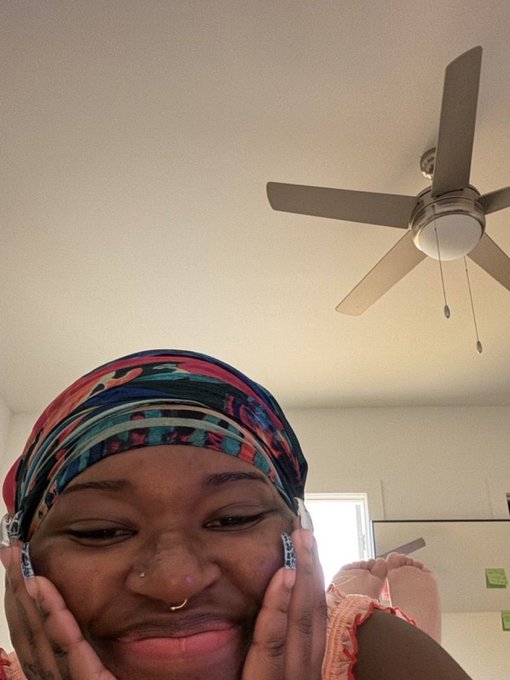 A woman with a colorful patterned headwrap rests her chin on her hands with a gentle, knowing smile, her nose ring and long decorated nails visible beneath a spinning ceiling fan. The contemplative, "been there" expression perfectly mirrors the post's playful jab at crypto holders who say they've quit but keep coming back.