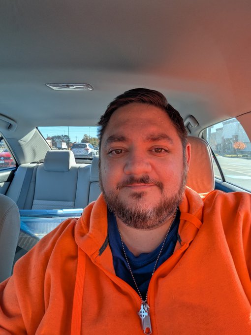 A bearded man in an orange hoodie takes a selfie inside a car, with daylight, traffic, and buildings visible through the windows.