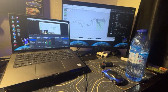 A trading or streaming setup features a laptop screen showing OBS studio and a separate monitor displaying financial charts, with a water bottle, a small toy car, and other items on the desk.