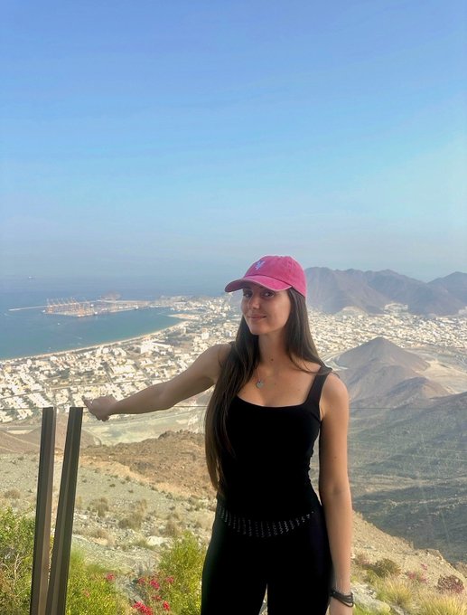 A woman wearing a pink cap and black tank top stands at a scenic overlook, with a panoramic view of a coastal city, mountains, and ocean behind her.