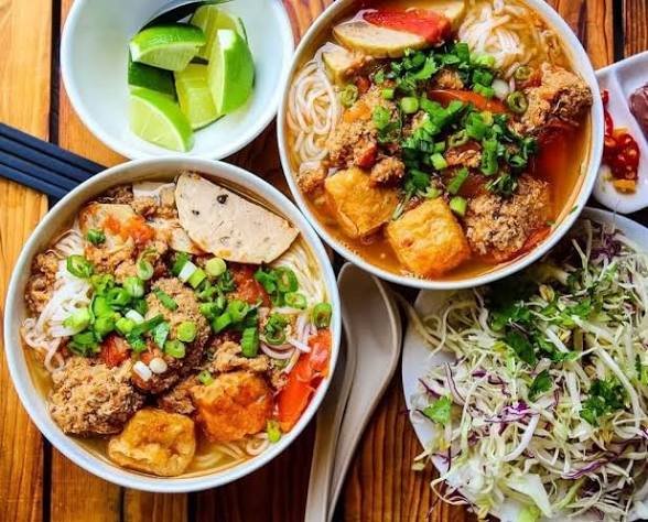 An overhead view of a meal featuring two bowls of noodle soup topped with fried tofu, meat, green onions, and lime wedges, served with a side of shredded cabbage salad.