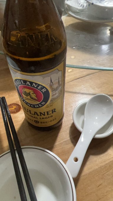An empty Paulaner München Original Lager beer bottle sits on a wooden table alongside a white soup spoon on a saucer, chopsticks, and the edge of a bowl in what appears to be a restaurant setting.