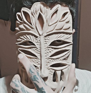 A person with tattooed fingers holds an intricate carved wooden mask featuring tree-like cutout designs over their face.