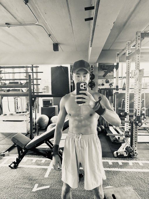 A black-and-white photo of a shirtless man in a gym taking a mirror selfie, wearing shorts and a cap, with workout equipment visible behind him.