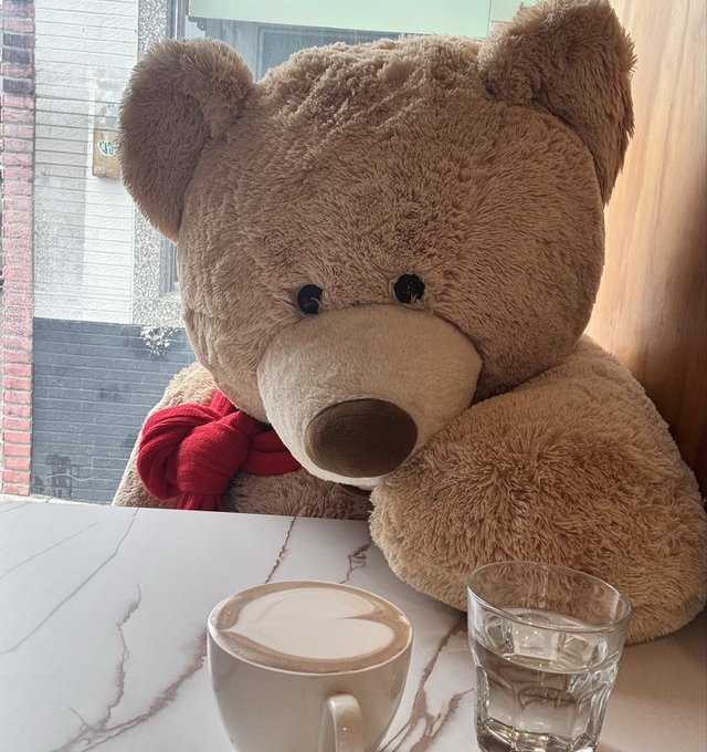 A large brown teddy bear with a red bow sits behind a marble table holding a cappuccino with latte art and a glass of water.