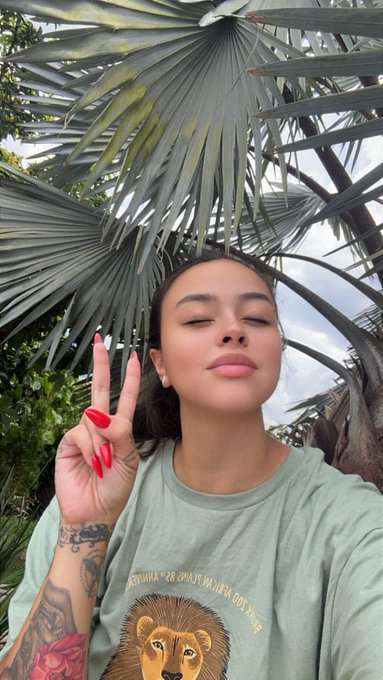 A woman with red nail polish poses for a selfie making a peace sign under a fan palm, wearing a green t-shirt featuring a lion graphic.