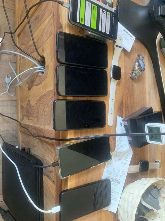 Multiple smartphones lined up on a wooden table all charging simultaneously, accompanied by an Apple Watch, a FitBit-style watch, and various receipts and cables scattered around.