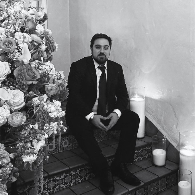A black-and-white photograph shows a man in a dark suit and tie sitting on tiled steps next to large floral arrangements and pillar candles.