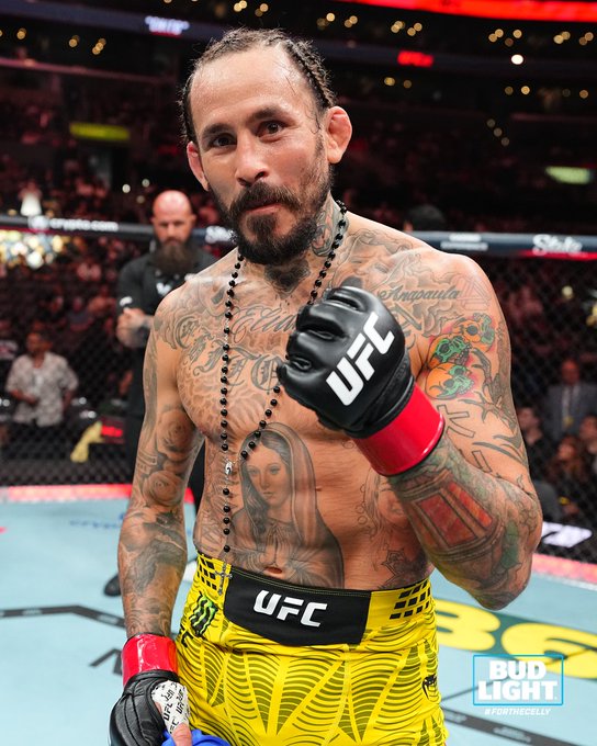 A heavily tattooed UFC fighter wearing yellow shorts and black UFC gloves stands in the octagon with a rosary around his neck, posing with a raised fist after a victory at UFC 331. The "Bud Light" logo is watermarked in the corner, aligning with the post's branding promotion as the fighter celebrates in the LA prelims event.