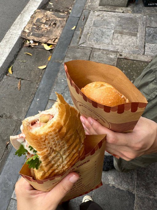 Two hands hold food items wrapped in paper on a sidewalk: a flatbread wrap and a bread roll.