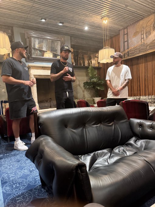 Three men in casual streetwear stand in a lounge featuring dark leather furniture, a fireplace, and ornate decor, with one man in a black cap speaking while the others listen.