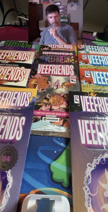 The image features a man sitting at a table with several books spread out around him. The books are of various sizes, indicating that he has purchased multiple copies of the comic series "VeeFriends" from @veefriends. The man is drinking something while looking at the books, possibly enjoying his new collection or preparing to read them. The post about the comics received by the community member highlights the positive and supportive nature of the @veefriends community, as they have gifted him a third comic book in addition to the two he already has.