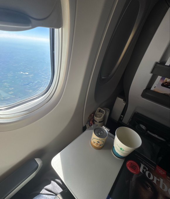 An airplane window view shows the sky and land below, with a fold-down tray holding a paper cup, a small bottle, and a Forbes magazine in the seatback pocket.