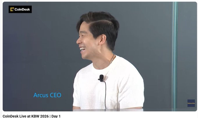 A screenshot from CoinDesk Live at KBW 2026 Day 1 showing the Arcus CEO, an Asian man in a white shirt with a lapel microphone, laughing in profile view.