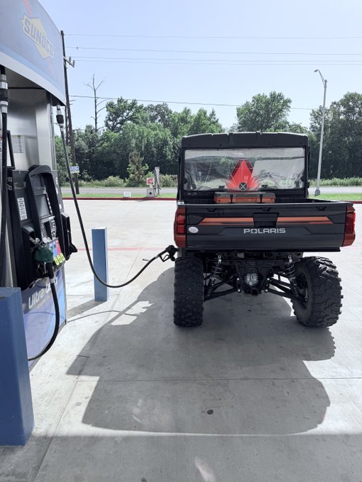 A Polaris UTV is parked at a fuel station with a nozzle inserted into its tank, with a red gas can visible in the bed of the vehicle, capturing a routine moment of country life maintenance.
