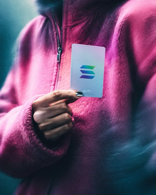 A person in a bright pink fleece jacket holding a translucent card with a blue and green logo.