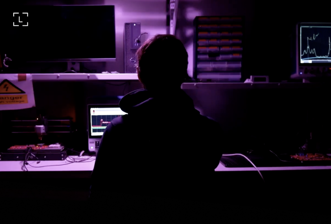 A silhouetted figure sits at a dimly lit workstation bathed in purple light, surrounded by multiple monitors—one displaying graphs and charts—evoking the shadowy world of cyber intrusions. A small "cs" target icon hints at vulnerability, reinforcing the caption's warning that AI agents can be exploited to drain funds even without malicious intent.