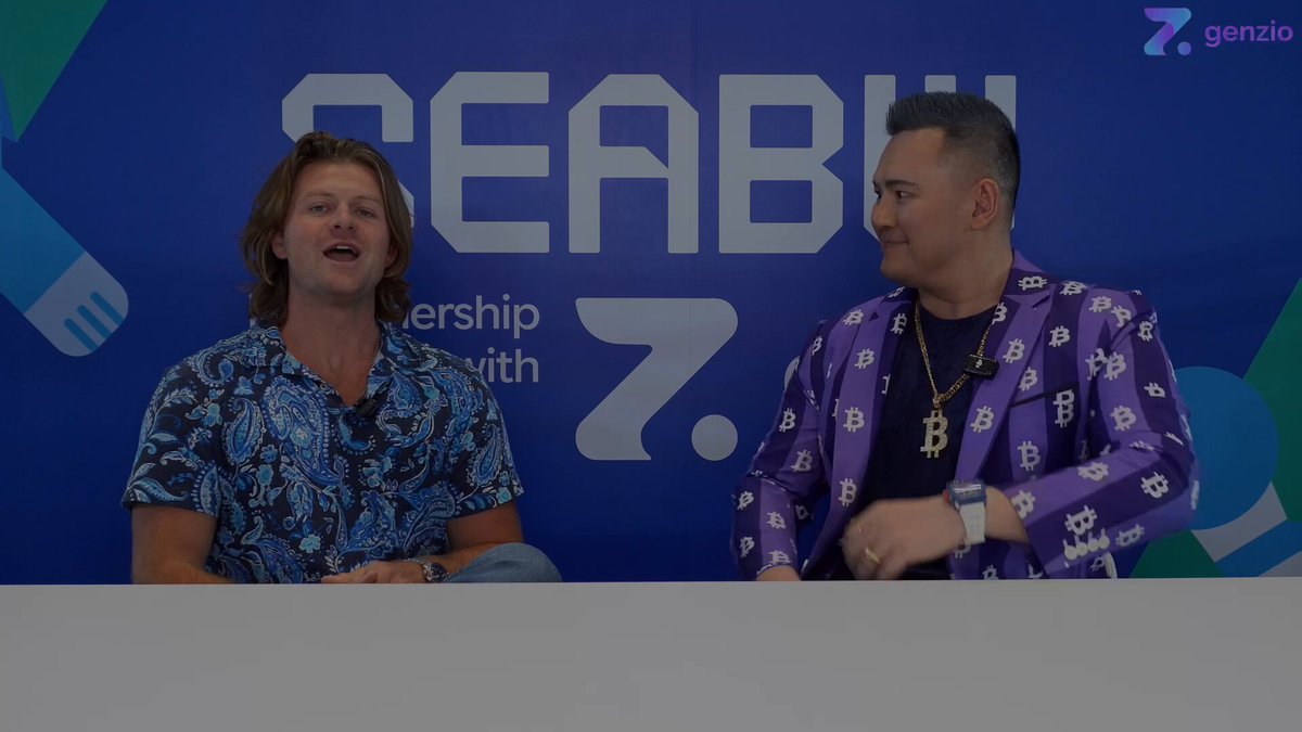 The image shows two men sitting at a table, talking about Bitcoin. One man is wearing a purple jacket with a patterned design, while the other has a blue shirt. They are both smiling as they engage in conversation. The background features a large blue banner that says "SEABU", and there's also a logo for "Genio" on the wall behind them.