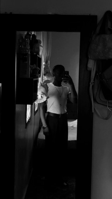 The image shows an individual holding up their phone with a mirror in front of them while taking a photo. The photograph has a vintage aesthetic, which adds character to the scene. In addition to the person taking the picture, there's also someone visible at the back of the frame who appears to be looking towards the camera or the reflection on the mirror screen. Another person is partially visible behind this individual but is not the main focus in the image. The overall atmosphere seems casual and relaxed as the person captures their morning moments through a self-portrait with a vintage feel.